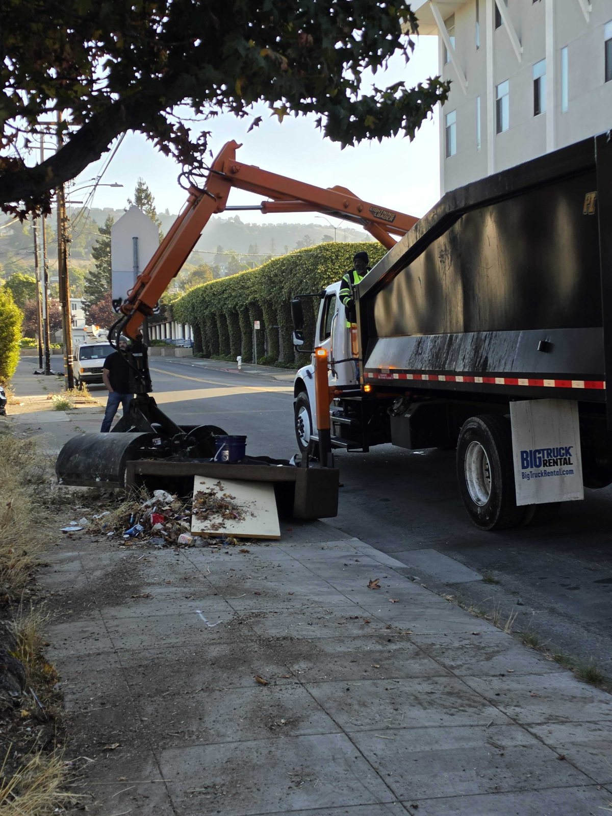 A lightning loader lifting a couch, a table, and various furniture off an Oakland sidewalk into its bed