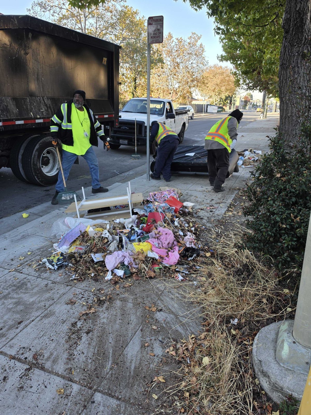 Three crew members in high-visibility vests working around a mixed pile of discarded furniture, clothing, and bags on a sidewalk